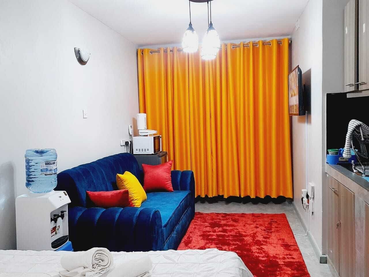 A compact room with a blue sofa, red and yellow cushions, a red rug, a water dispenser, a microwave, and a TV on a cabinet.