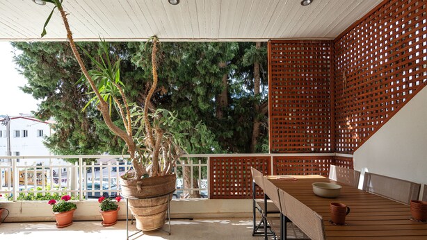 A balcony with a wooden table, chairs, potted plants, and a view of the city.