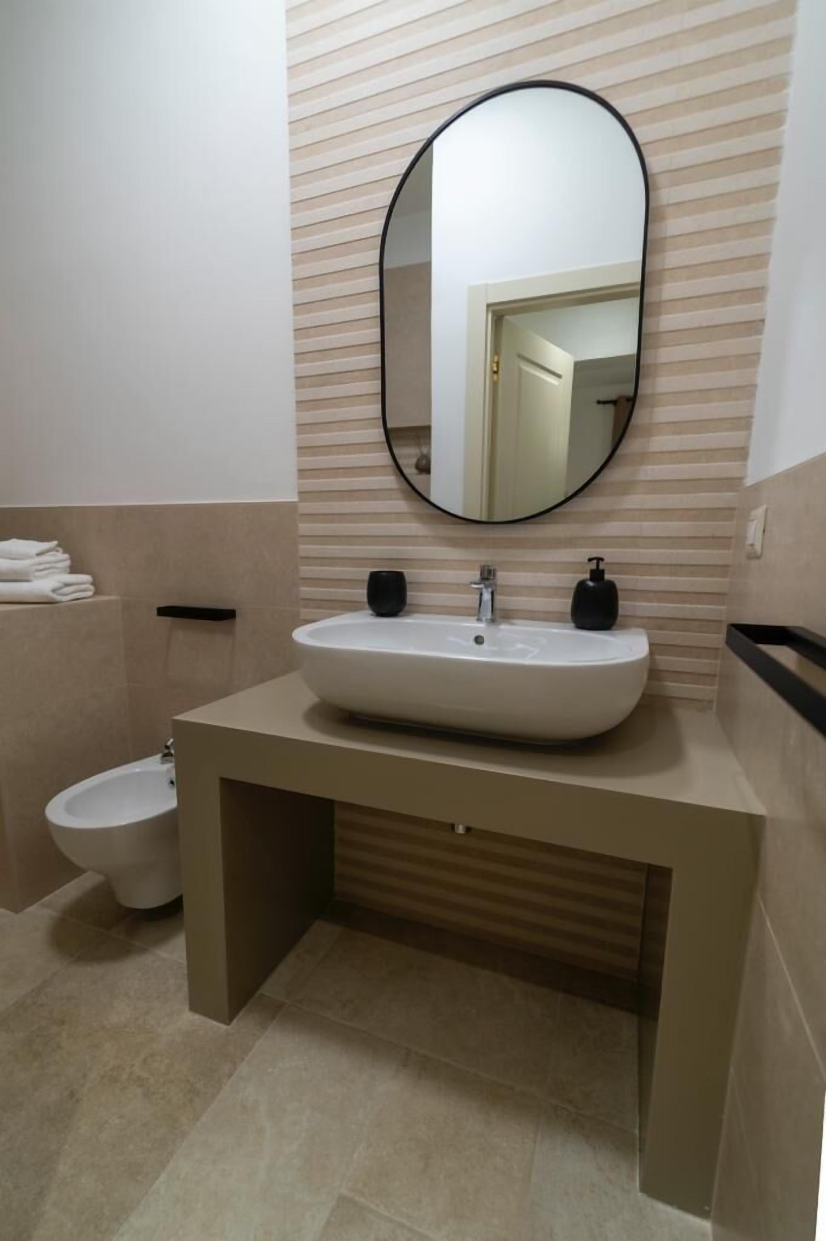 Une salle de bain moderne avec un grand miroir ovale, un lavabo blanc, un distributeur de savon noir et des toilettes.