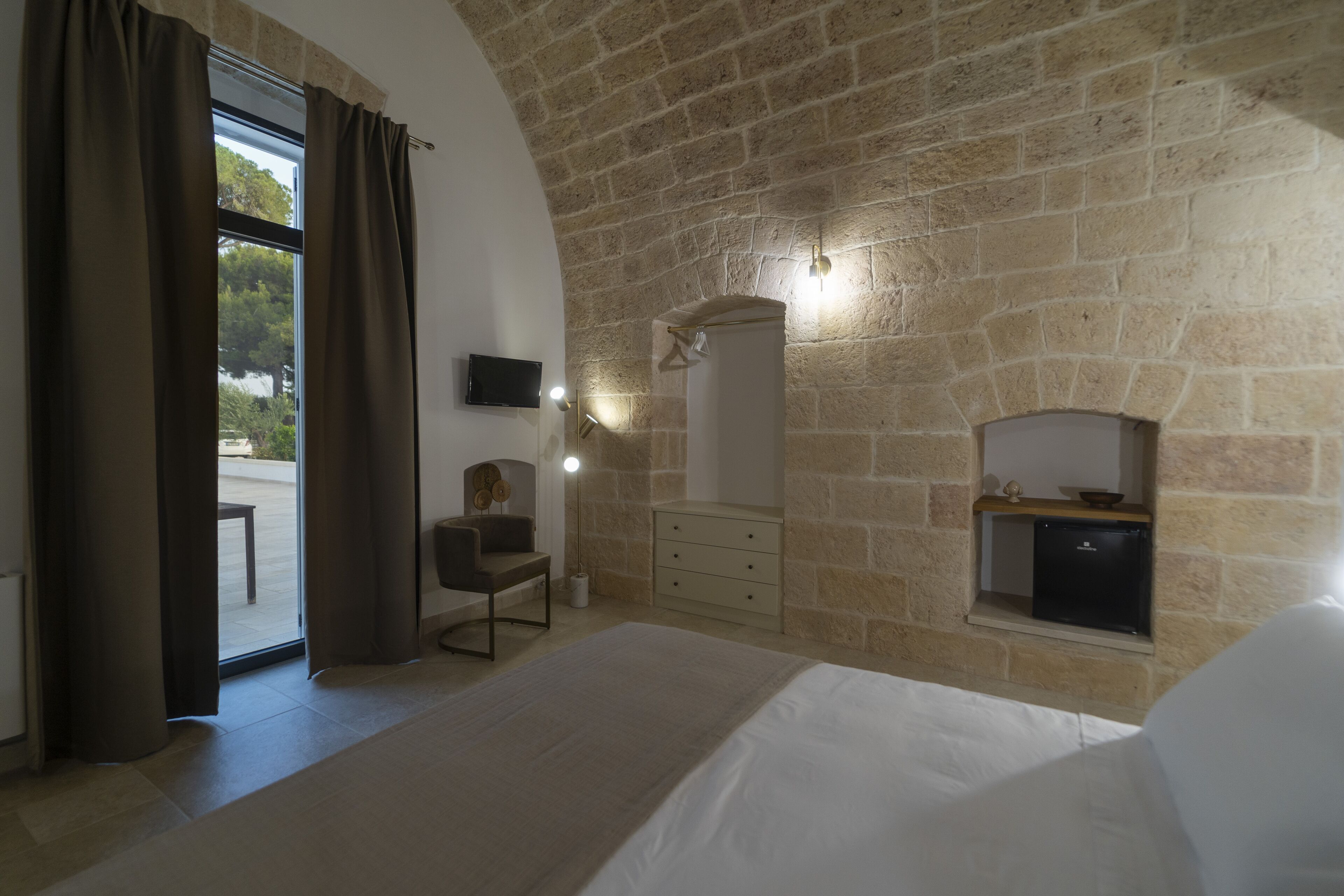 A bedroom with a stone wall, a bed, a chair, a dresser, a TV, and a window with curtains.
