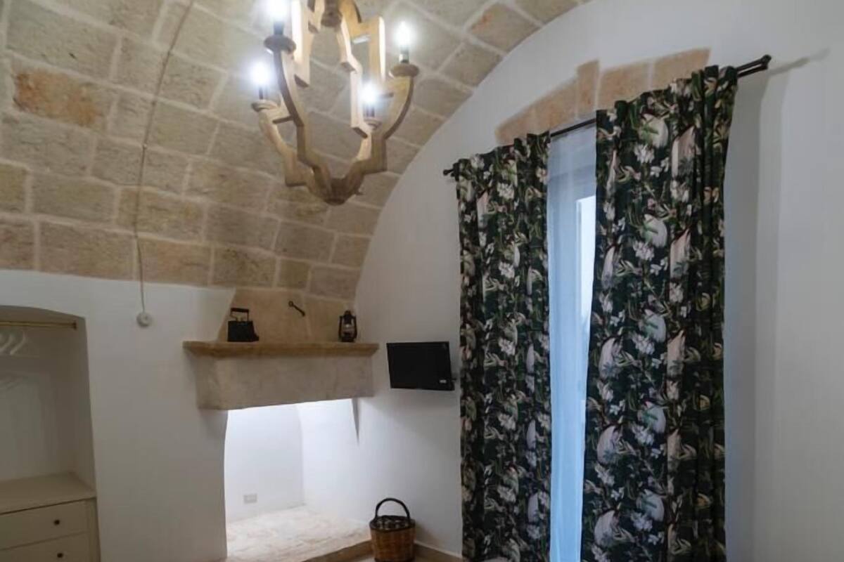 A bedroom with a stone ceiling, a wooden bed, a small shelf with a kettle, a television, and floral curtains.