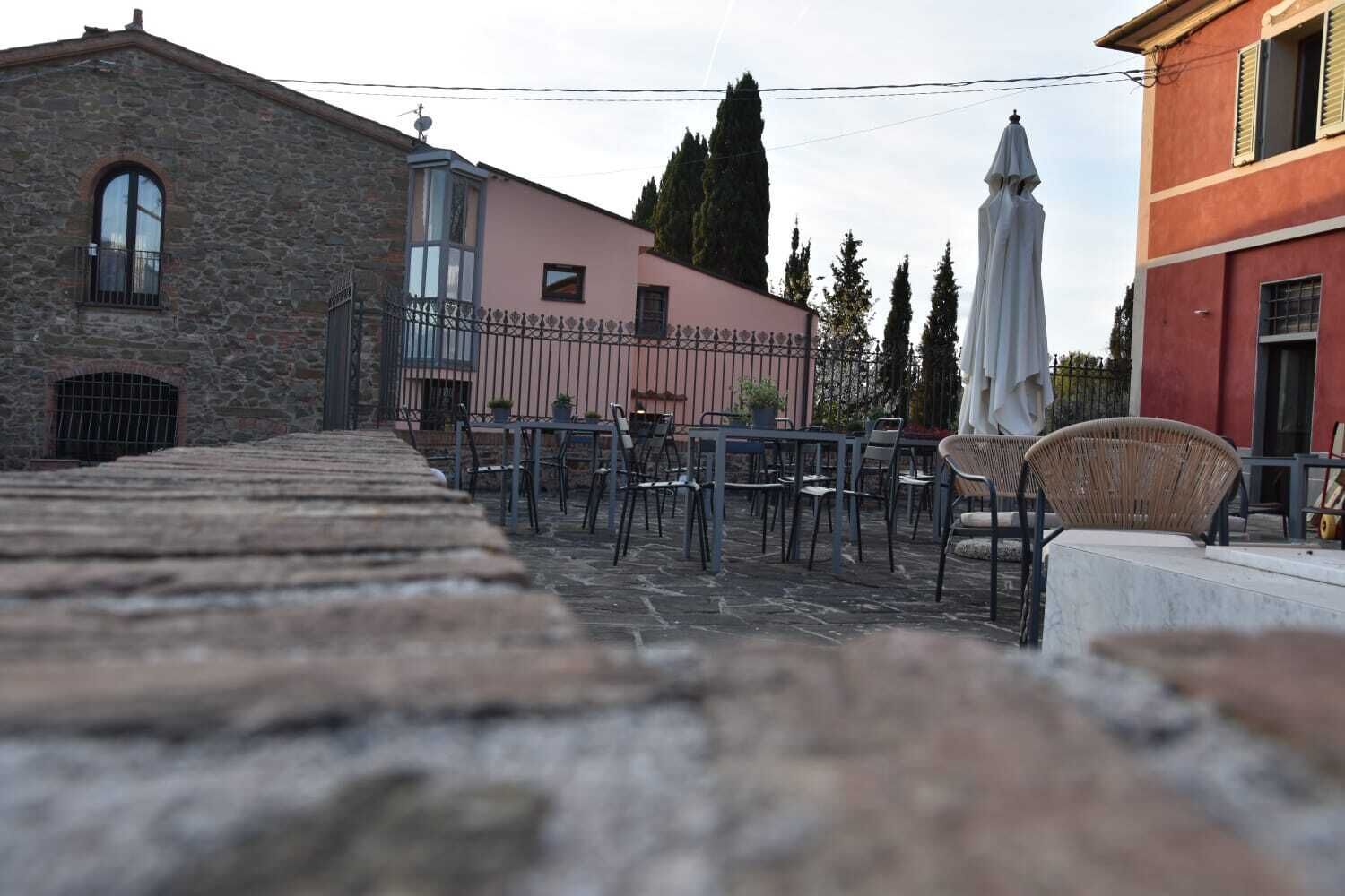 Un área de estar al aire libre con suelo de piedra, mesas y sillas de metal, rodeada de edificios con fachadas de piedra y pintadas.