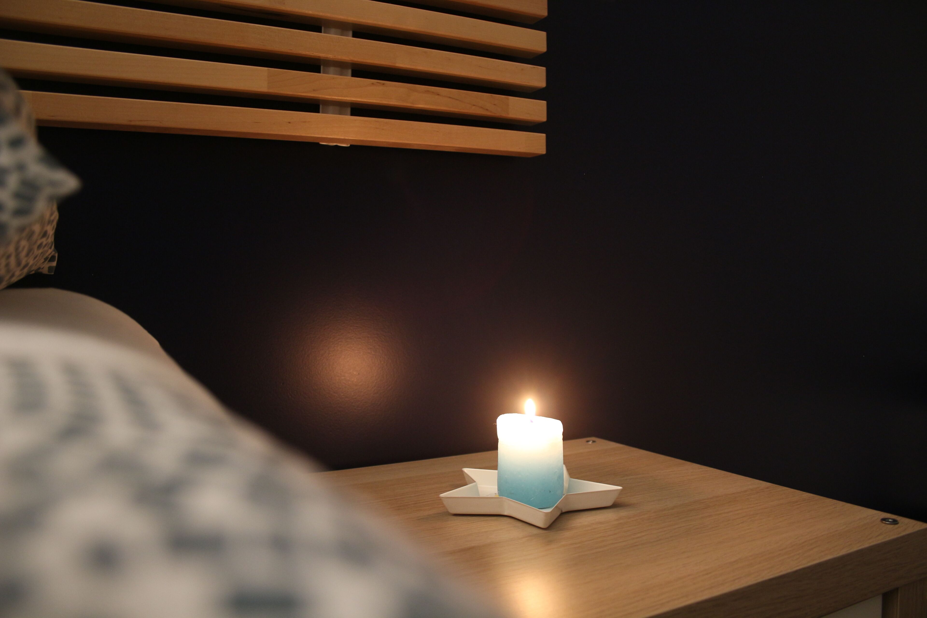 A bedside table with a lit candle, a wooden headboard, and a dark wall.