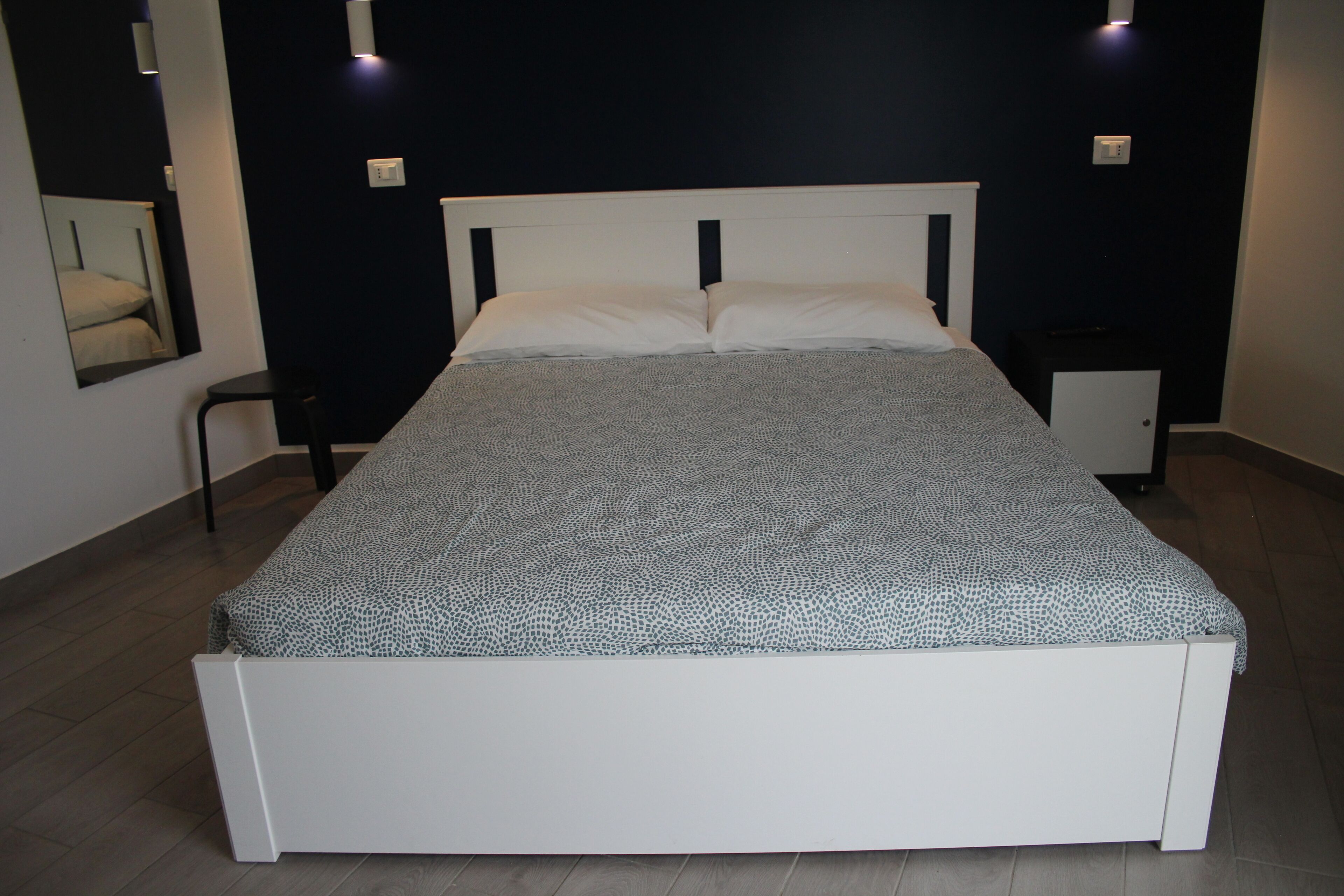 Una cama con cabecera blanca y colcha estampada en una habitación con paredes oscuras y una mesita de noche pequeña.