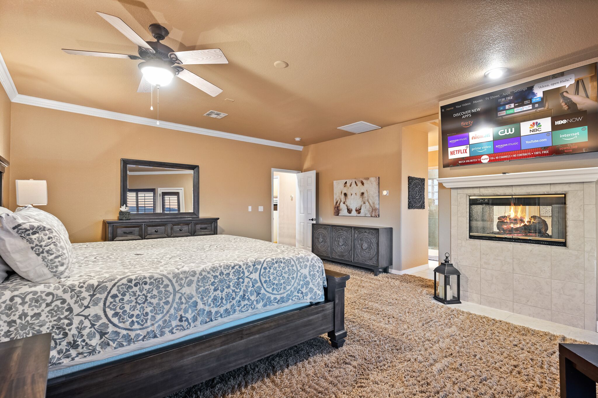 A bedroom with a bed, a ceiling fan, a fireplace, and a mounted TV displaying various channels.