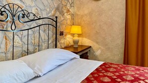 Un lit avec du linge de lit blanc, une couvre-lit rouge à motifs, une tête de lit en métal, un mur à la texture rappelant la pierre, une petite table avec une lampe jaune et une assiette décorative fixée au mur.