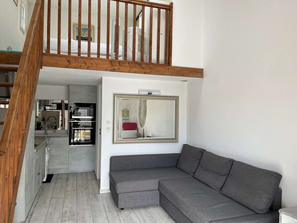 A modern loft apartment with a wooden staircase, a grey sectional sofa, and a kitchen area visible through an open door. - Maison 2 pièces 4 couchages PORT LEUCATE NATURISME (PORT LEUCATE NATURISME)