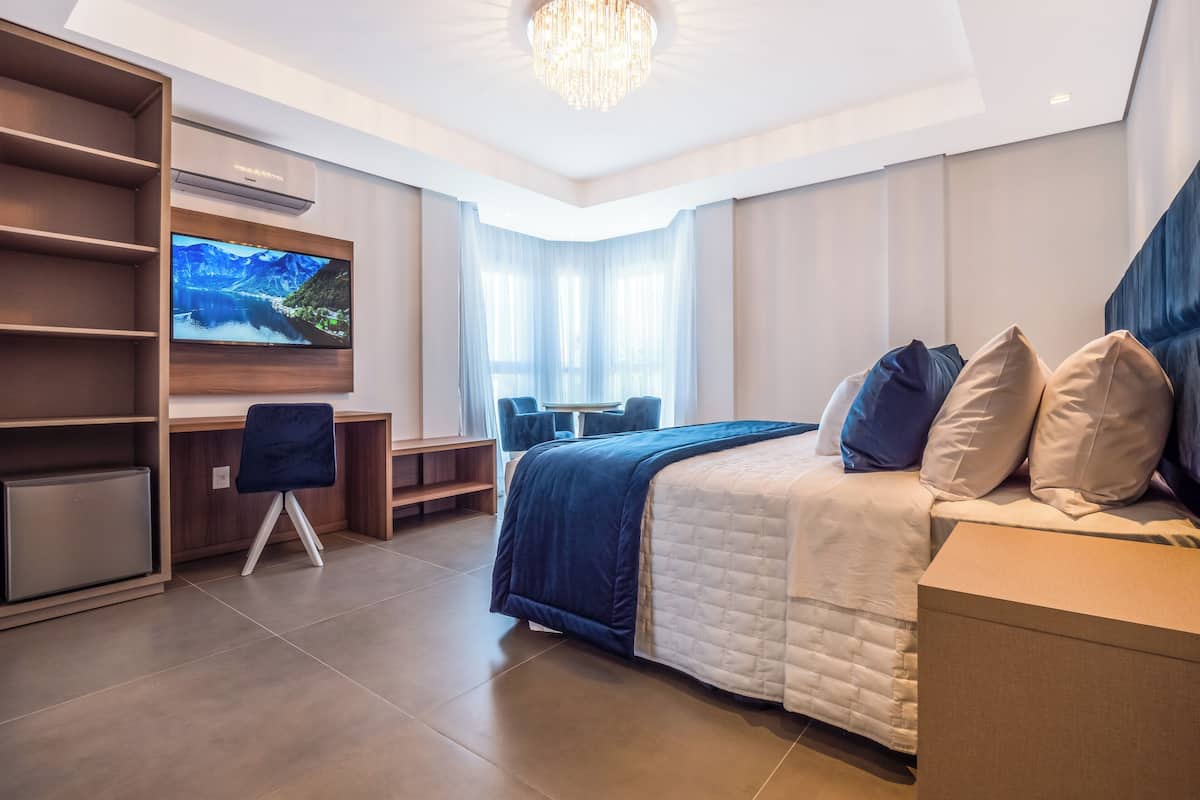 A modern hotel room with a large bed, a desk with a chair, a TV mounted on the wall, and a chandelier.