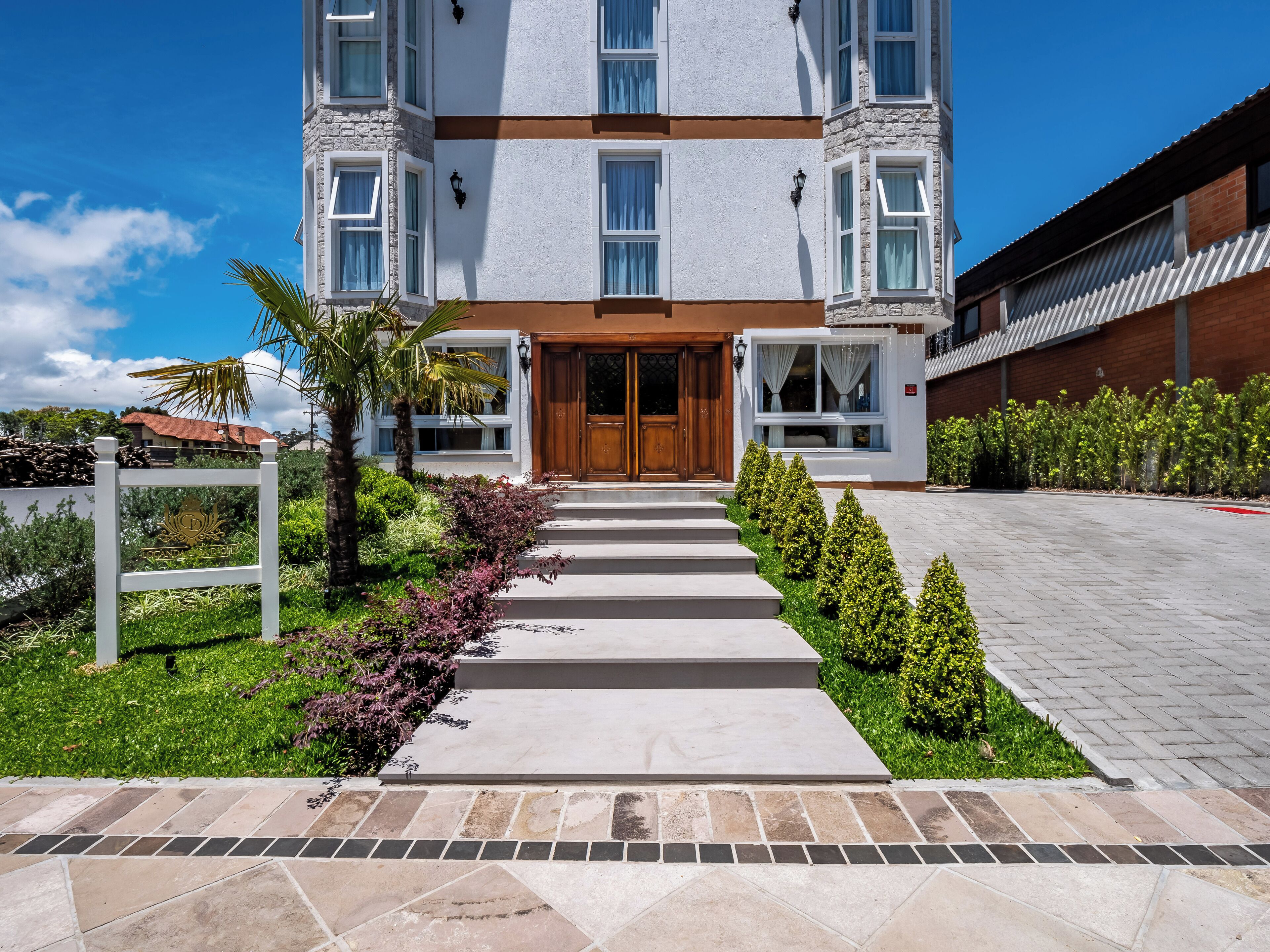 Um hotel moderno com entrada central, fachada de pedra e um jardim bem cuidado com um caminho que leva à entrada.