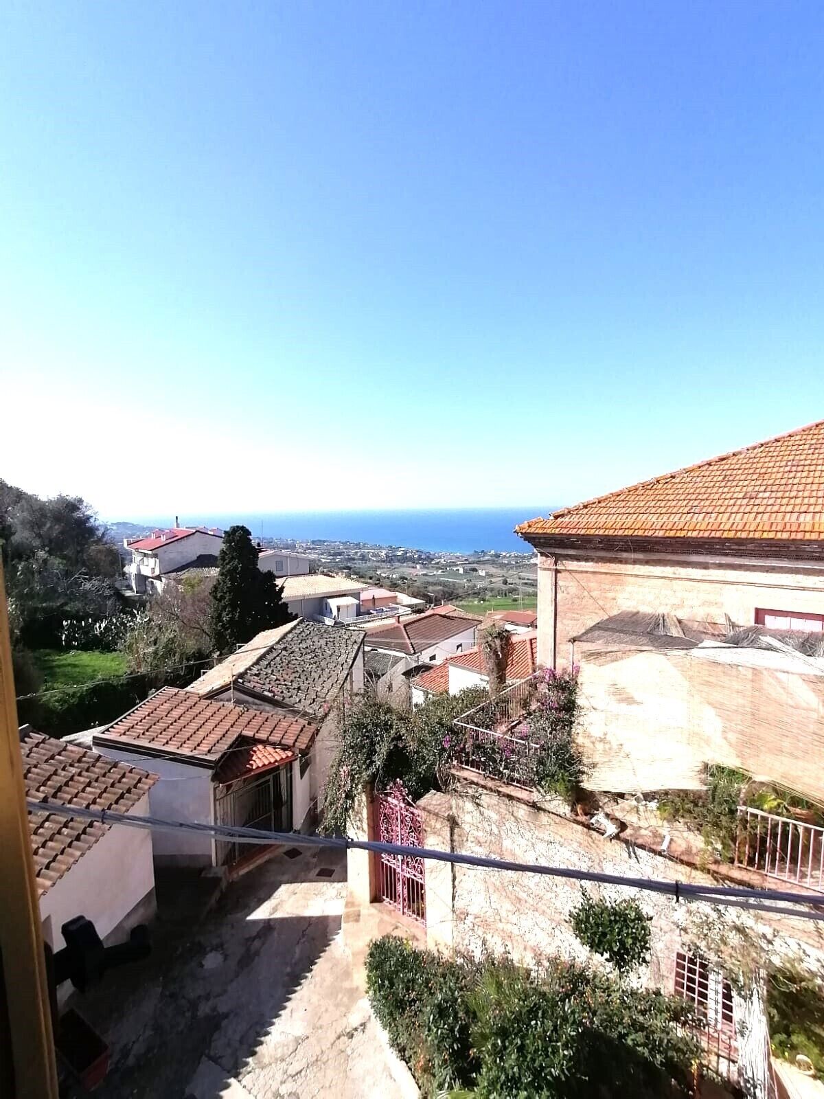A view of a residential area with terracotta-tiled roofs, a clear blue sky, and a distant view of the sea.