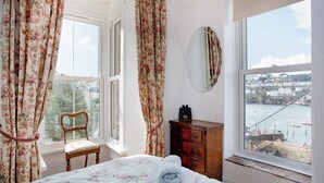 Ein Schlafzimmer mit einem Bett, einem Holzstuhl, einer Kommode und Blick auf einen Hafen durch ein großes Fenster.