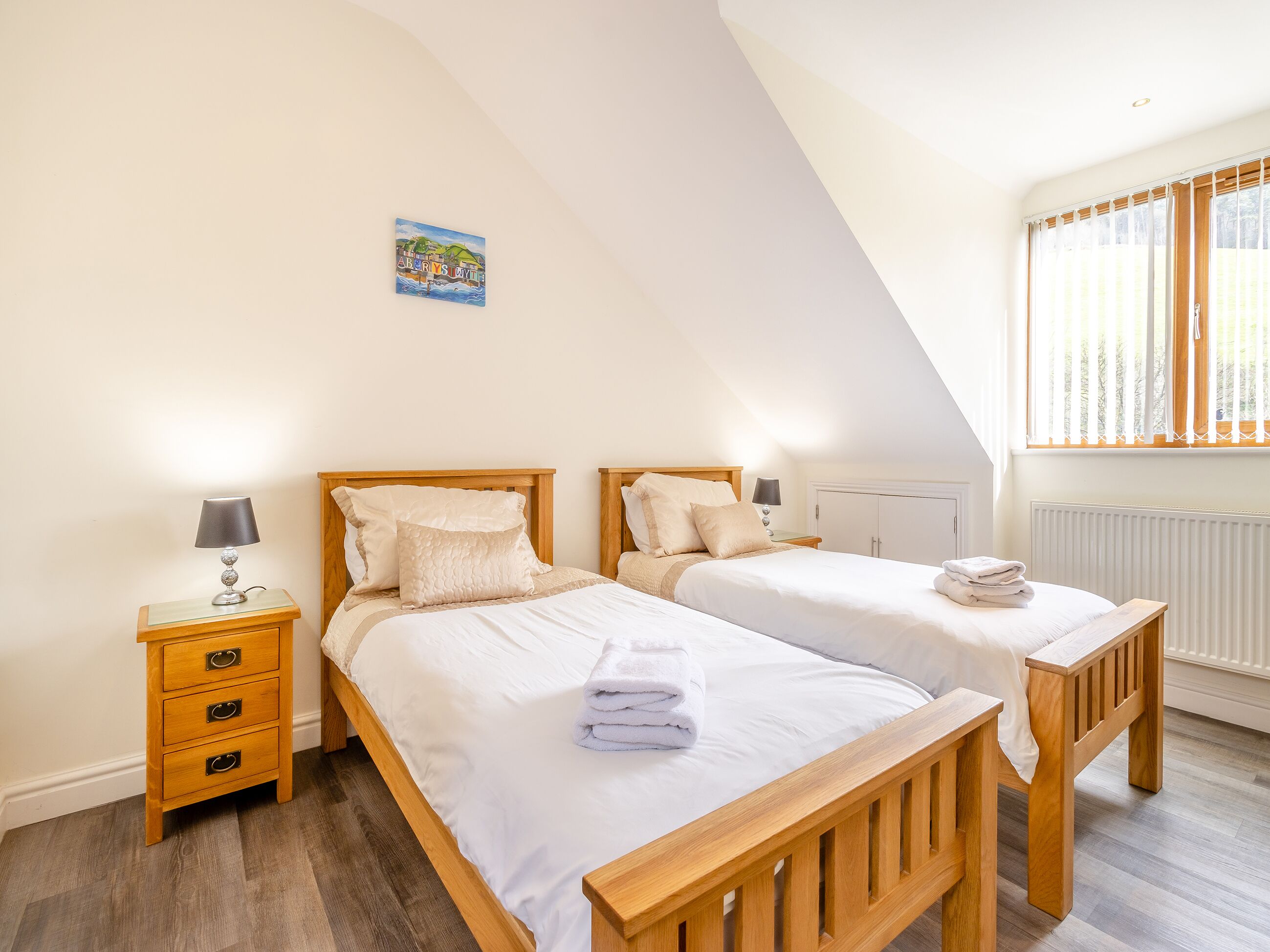A bedroom with two wooden beds, a nightstand, a lamp, a radiator, and a window with blinds.