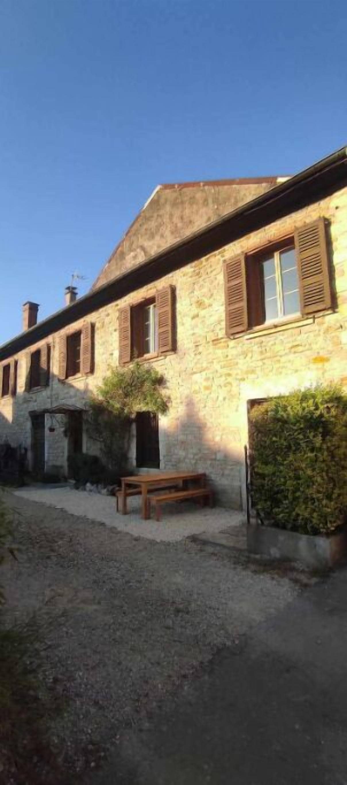 Un bâtiment en pierre avec une cour recouverte de gravier, un banc et un petit arbre.