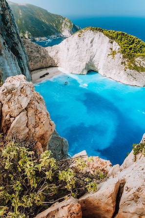 A coastal cliff with a clear blue sea, rocky terrain, and sparse vegetation.