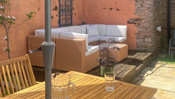 Une table en bois avec deux verres et une carafe, un canapé en osier avec des coussins blancs et une plante en pot.