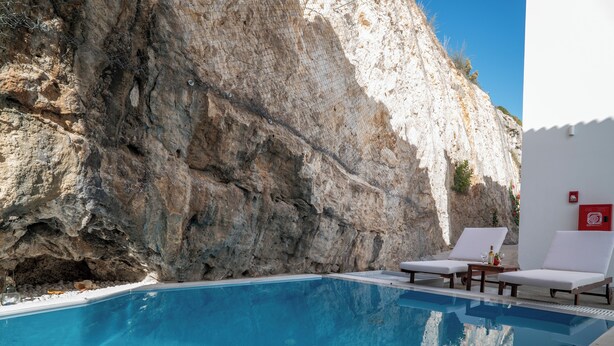 Una piscina con sillas de descanso blancas y un acantilado rocoso de fondo.