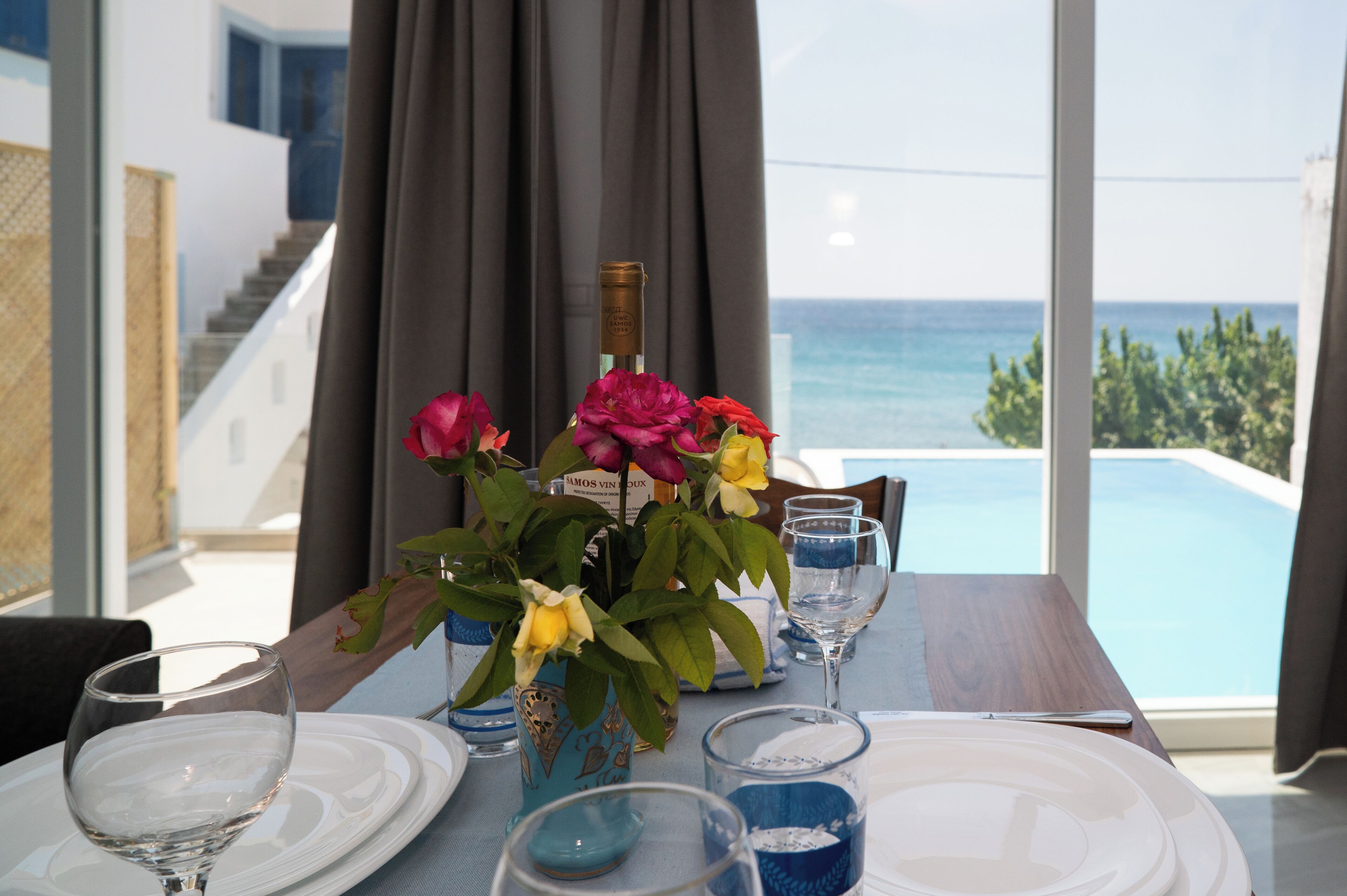 Une table à manger avec une bouteille de vin, un vase rempli de fleurs et une vue sur la mer par de grandes fenêtres.