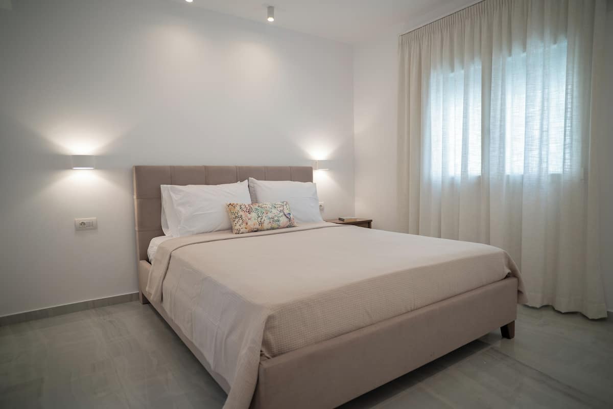 Un dormitorio moderno con una cama grande, dos lámparas de noche y una ventana con cortinas traslúcidas.