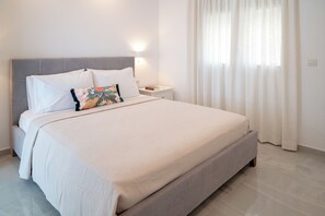 A neatly made bed with white linens and a grey headboard, a bedside table with a lamp, and a window with sheer curtains. - Kontemare Seascape (Samos)