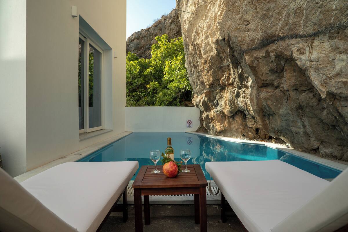 Zona junto a la piscina con dos sillones de descanso, una mesa con una botella y vasos, y vista a un risco y vegetación.