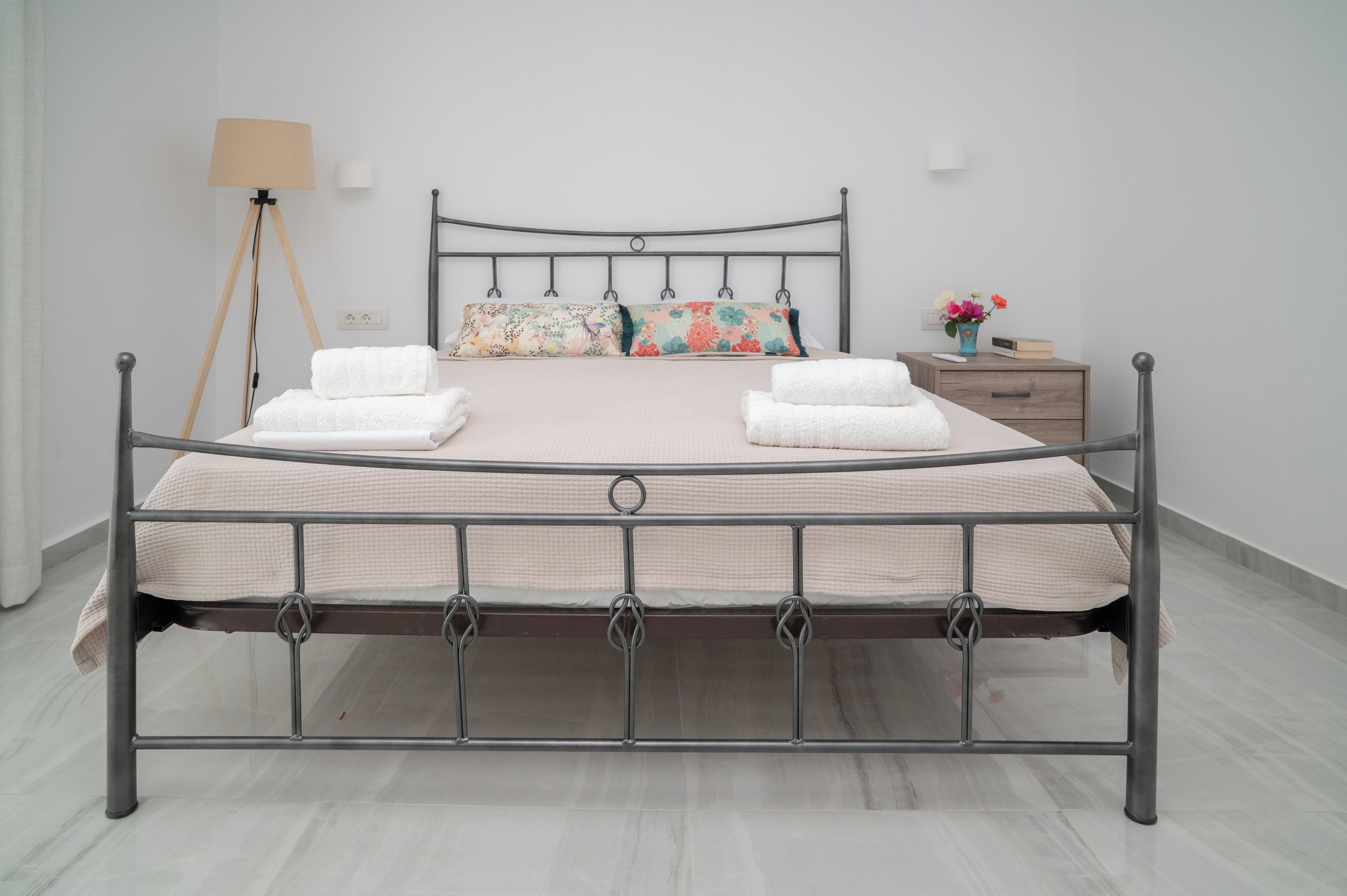 Una cama con estructura metálica, dos almohadas y dos toallas dobladas, una mesita de noche con un florero y una lámpara de pie.