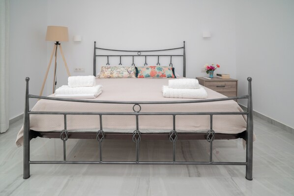 A metal-framed bed with two pillows and two folded towels, a bedside table with a vase of flowers, and a floor lamp.