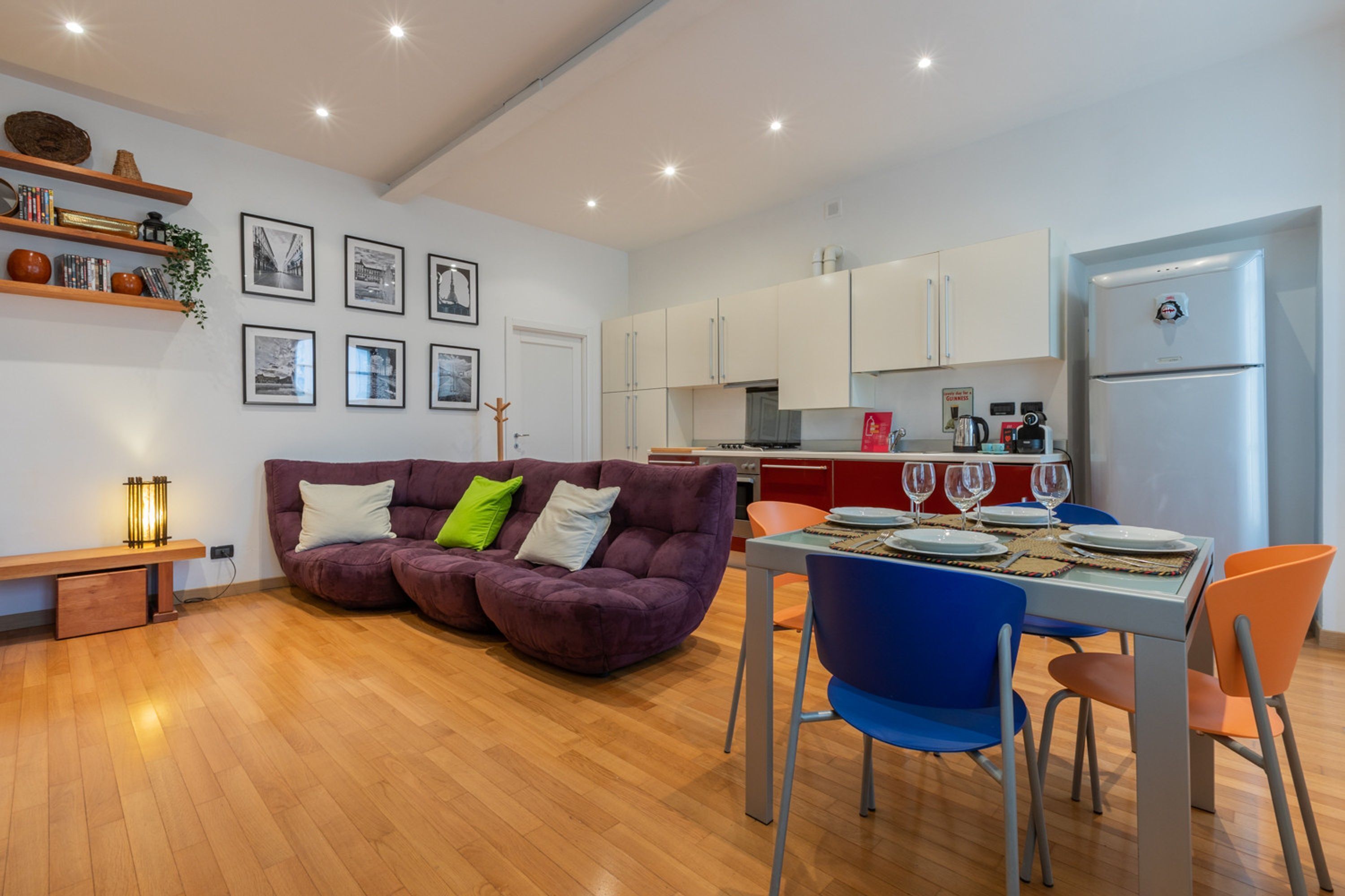 Een moderne keuken met witte kastjes, een inbouwoven en een koelkast. Er is een eethoek met een tafel gedekt voor twee personen, blauwe stoelen en een paarse bank met groen en witte kussens.