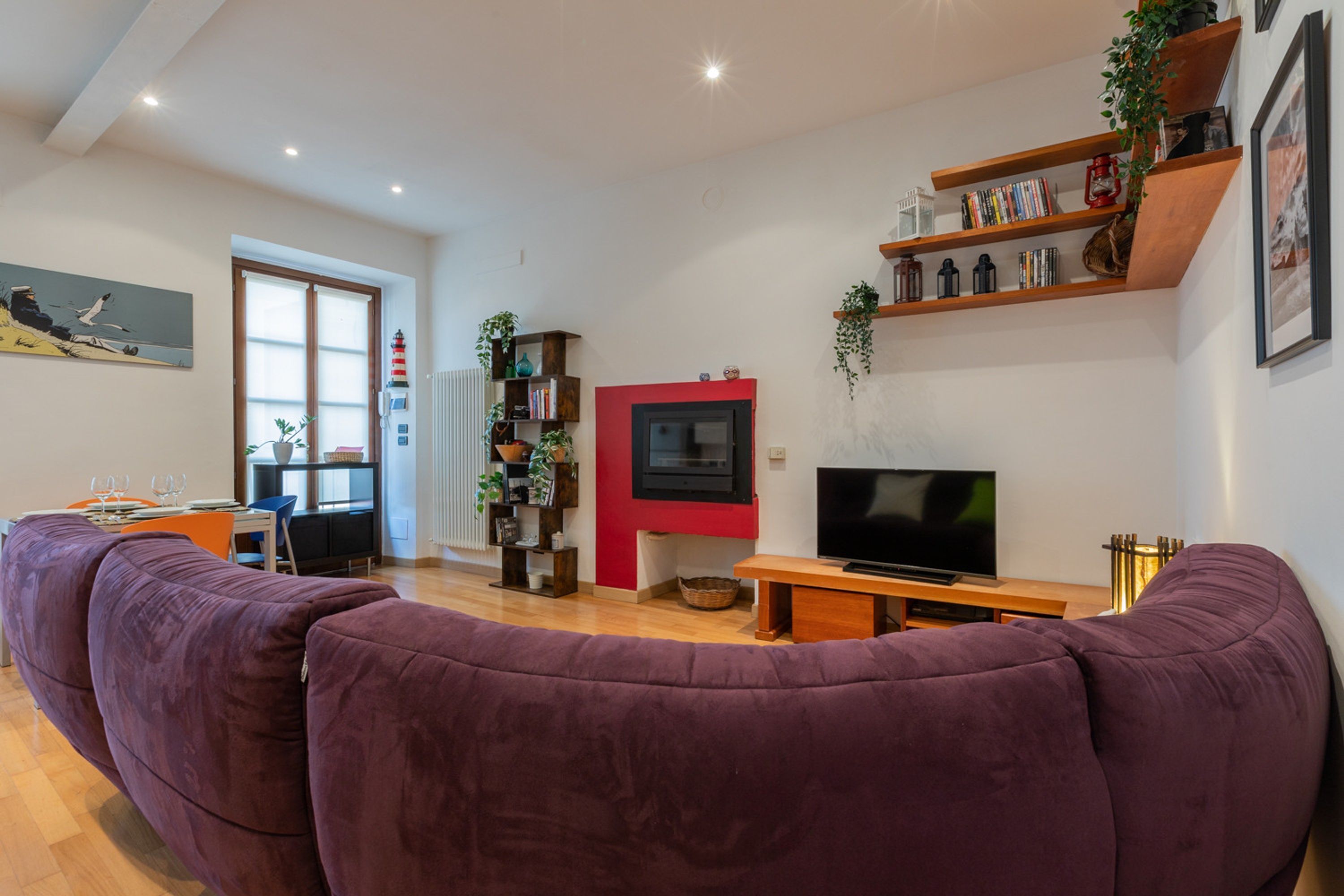 A living room with a large purple sofa, a TV, wooden shelves with books and plants, and a red accent wall.