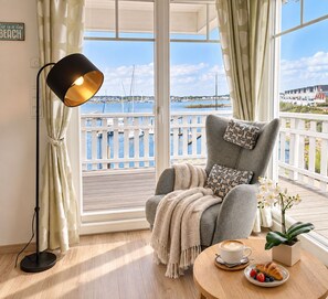 Ein Zimmer mit Blick auf eine Marina, ein grauer Sessel mit Überwurfdecke, eine Stehlampe und ein Tisch mit Kaffeetasse und Obst.