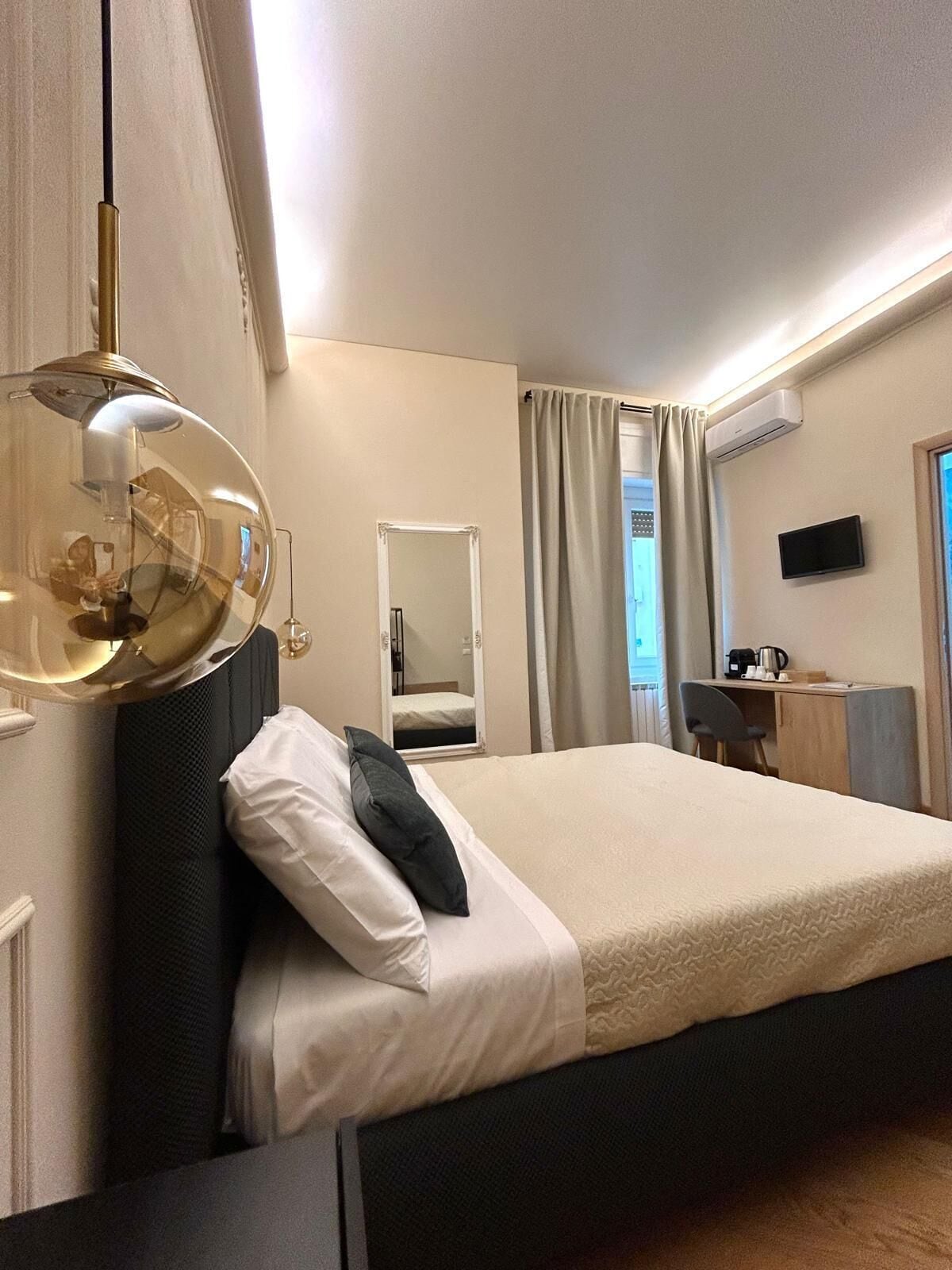 A hotel room with a large bed, a bedside table, a mirror, a TV, and an air conditioning unit.