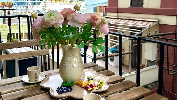 Un balcón con una mesa de madera, un jarrón con flores, una taza y una bandeja con aperitivos. Se ve un crucero al fondo.