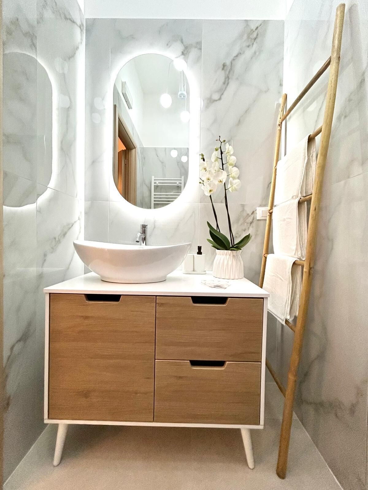 Un bagno con una parete in marmo, un lavandino bianco con un ampio specchio, cassetti in legno e una scala appoggiata al muro.