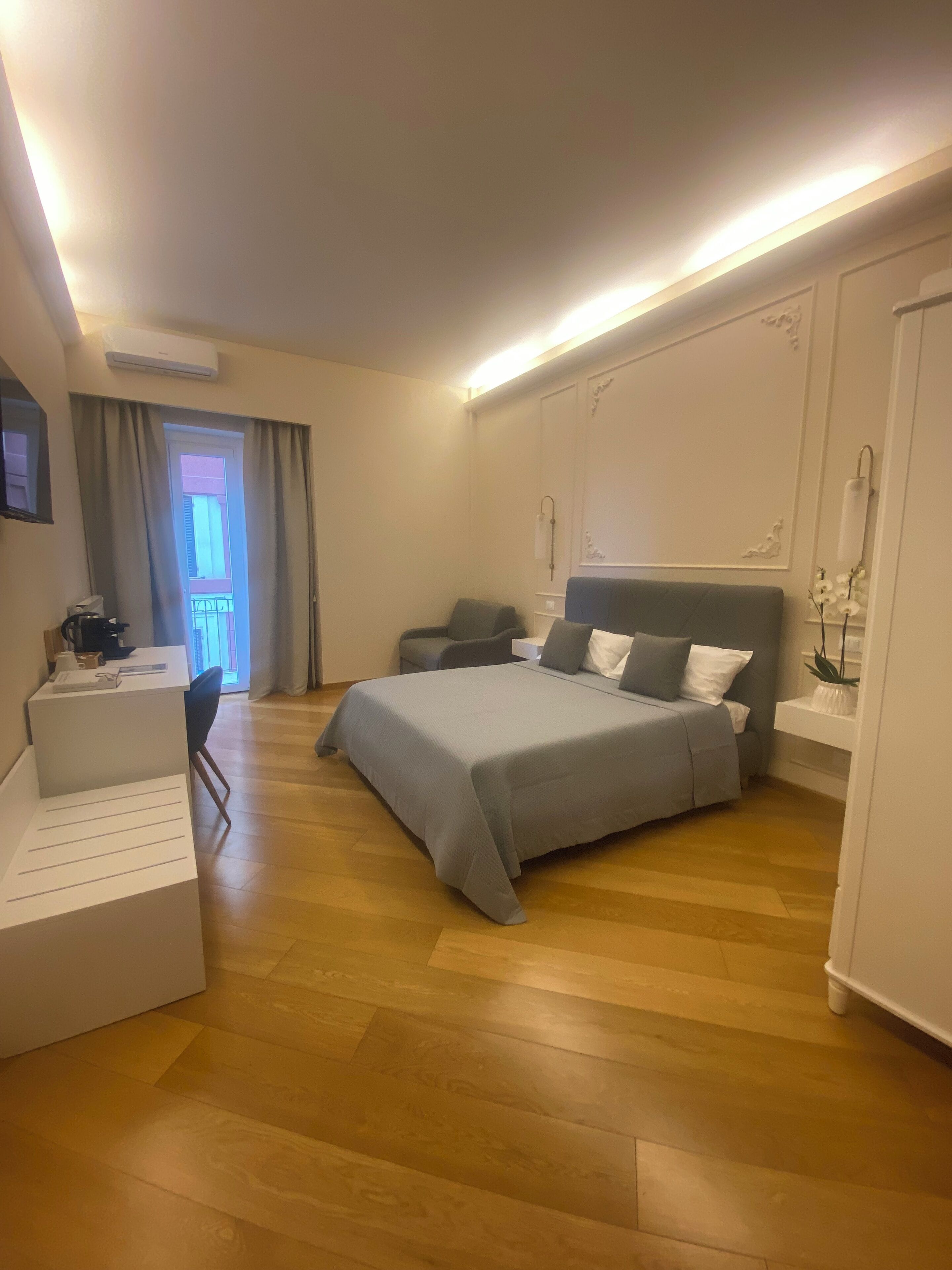 A modern hotel room with a large bed, a desk, and a chair. There is a balcony visible through the window.