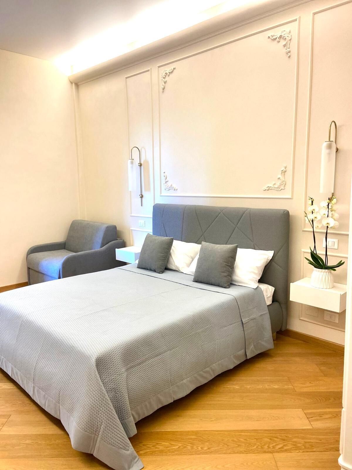 A modern bedroom with a large bed, a grey upholstered chair, and a bedside table with a lamp.