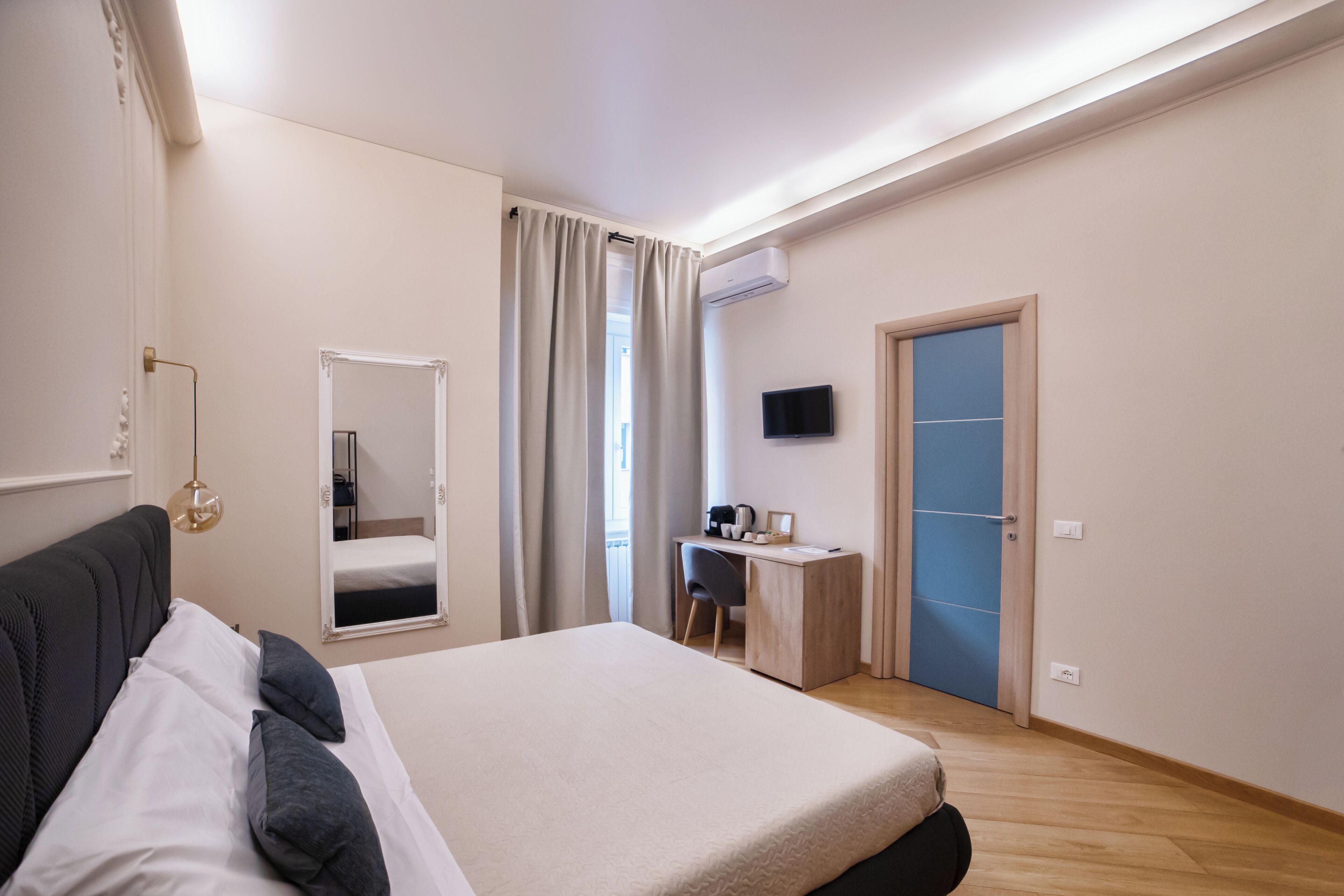 A modern hotel room with a large bed, a desk, a chair, a television, and a wardrobe.
