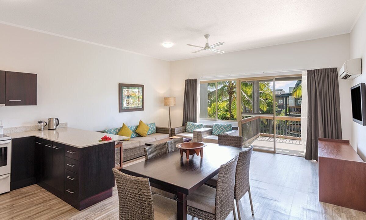 A modern kitchen with dark cabinetry, a dining area with wicker chairs, a sofa with patterned cushions, and a balcony with a view of palm trees.