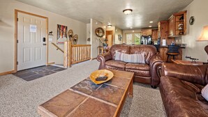 A spacious living room with a brown leather sofa, a coffee table with a remote control, and a kitchen area in the background. - Eden Mtn Retreat MH207 | Near Powder Mtn | Hot Tub (Eden)