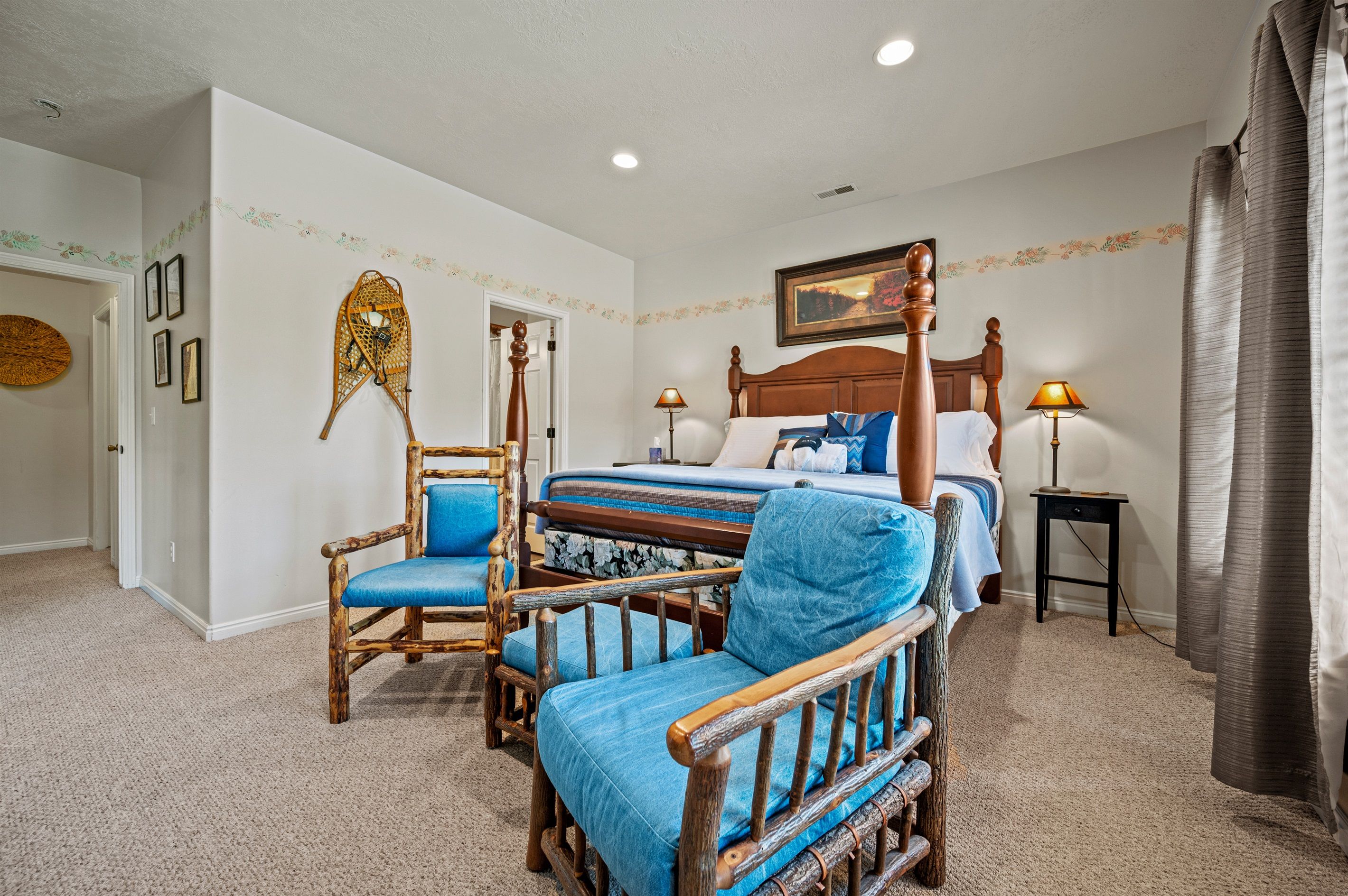 A bedroom with a wooden bed, two blue upholstered chairs, a small bedside table with a lamp, and a decorative wall hanging.