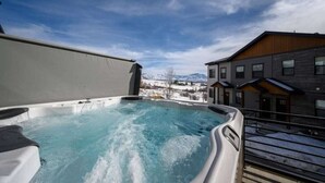 Un jacuzzi con vista a una montaña nevada, una terraza de madera y un edificio moderno con techo inclinado.