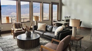 Ein geräumiges Wohnzimmer mit einer Ledersofa, Sesseln, einem Couchtisch und einem Essbereich mit Tisch und Stühlen. Große Fenster bieten Blick auf die Berge.