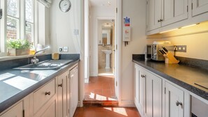 A narrow kitchen with white cabinets, a black countertop, and a window with blinds. There is a clock on the wall and a fire extinguisher mounted on the wall. - 3 bedroom accommodation in Overstrand, near Cromer (Overstrand, near Cromer)
