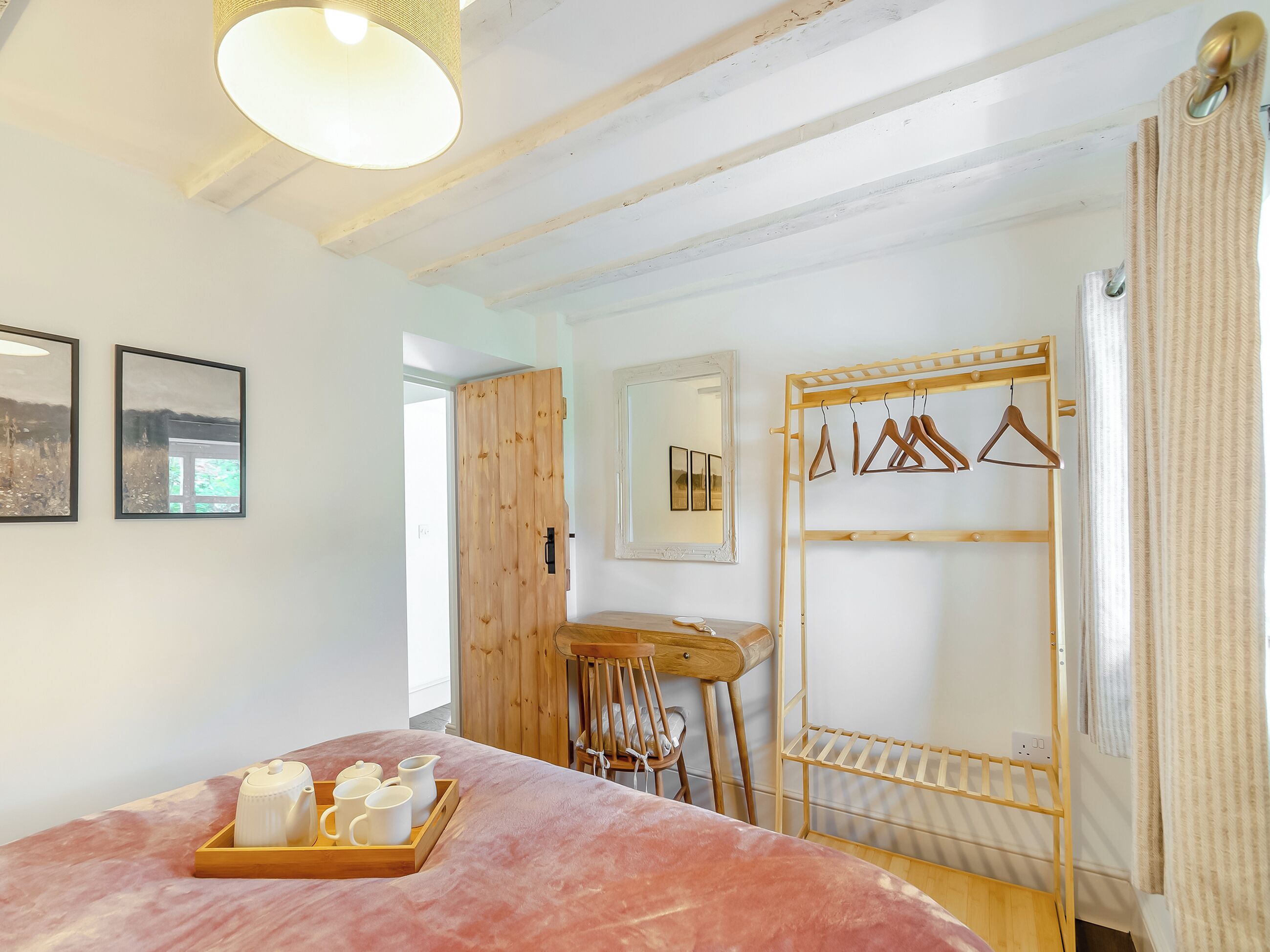 A bedroom with a bed, a wooden desk, a chair, a wooden coat rack, and a window with curtains.