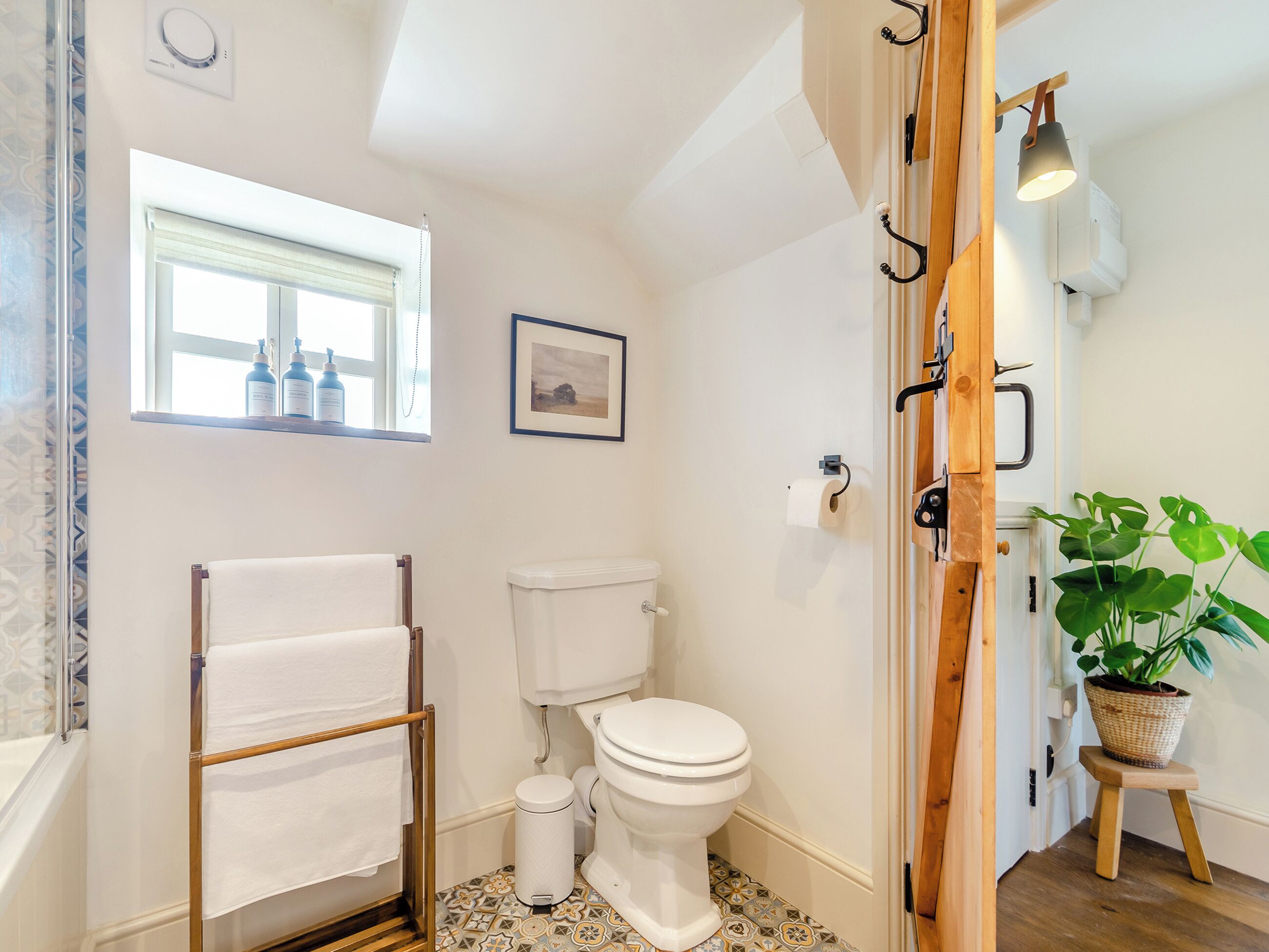 Ein Badezimmer mit Toilette, Handtuchhalter, Fenster mit Flaschen, einem gerahmten Bild und einem Blumentopf.