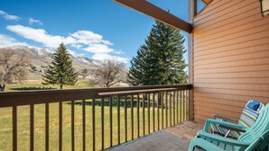 A balcony with two green Adirondack chairs, a wooden railing, and a view of a grassy field with trees and mountains in the background. - Sigi's Longhorn Lodge WL1409 |Gym |Ski Powder Mtn (Eden)