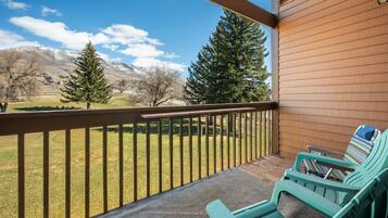 A balcony with two green Adirondack chairs, a wooden railing, and a view of a grassy field with trees and mountains in the background.