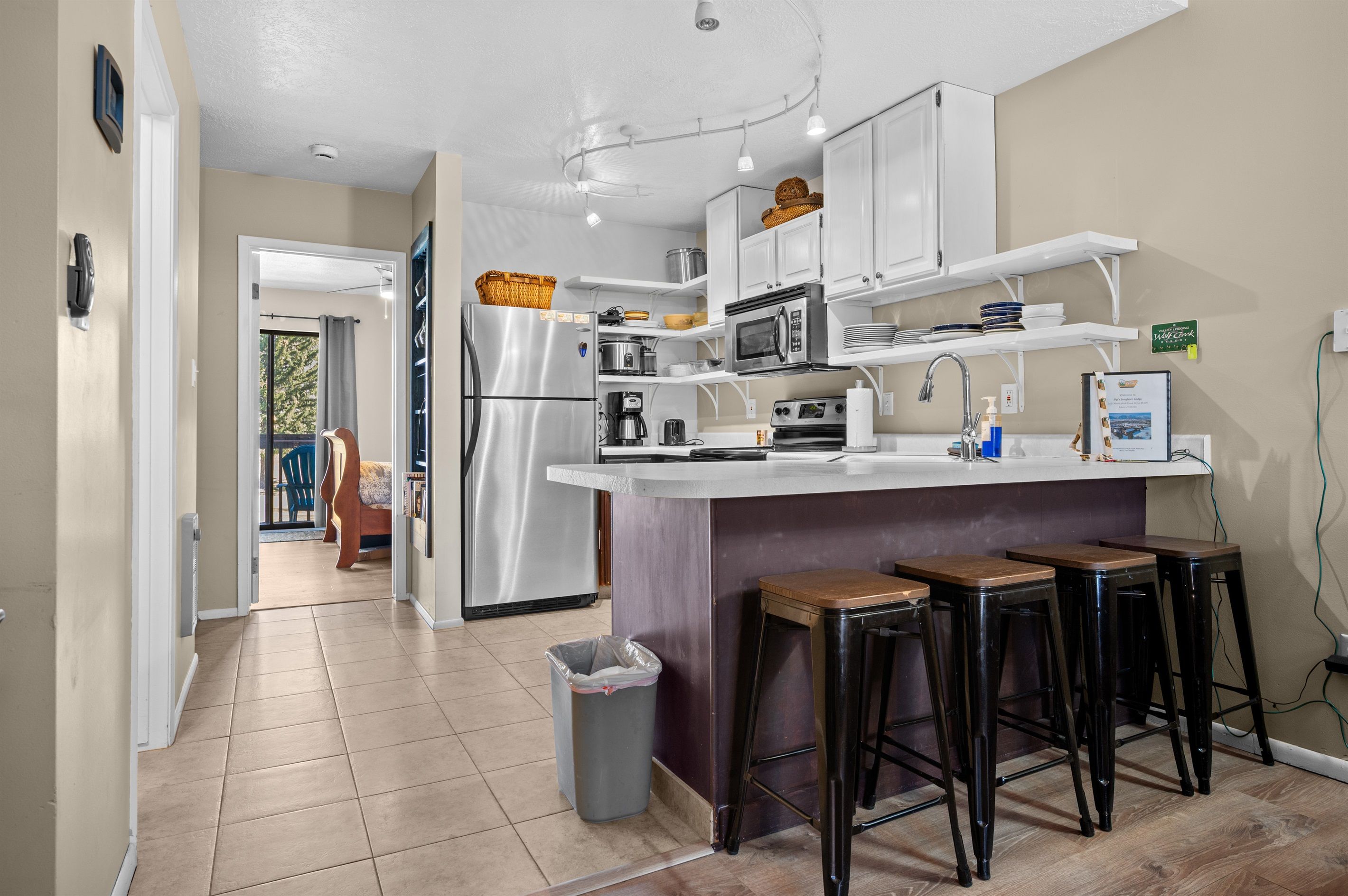 A modern kitchen with white cabinets, a stainless steel refrigerator, a microwave, and a coffee maker. There are three stools at the bar counter.