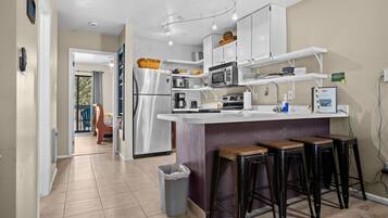 A modern kitchen with white cabinets, a stainless steel refrigerator, a microwave, and a coffee maker. There are three stools at the bar counter.