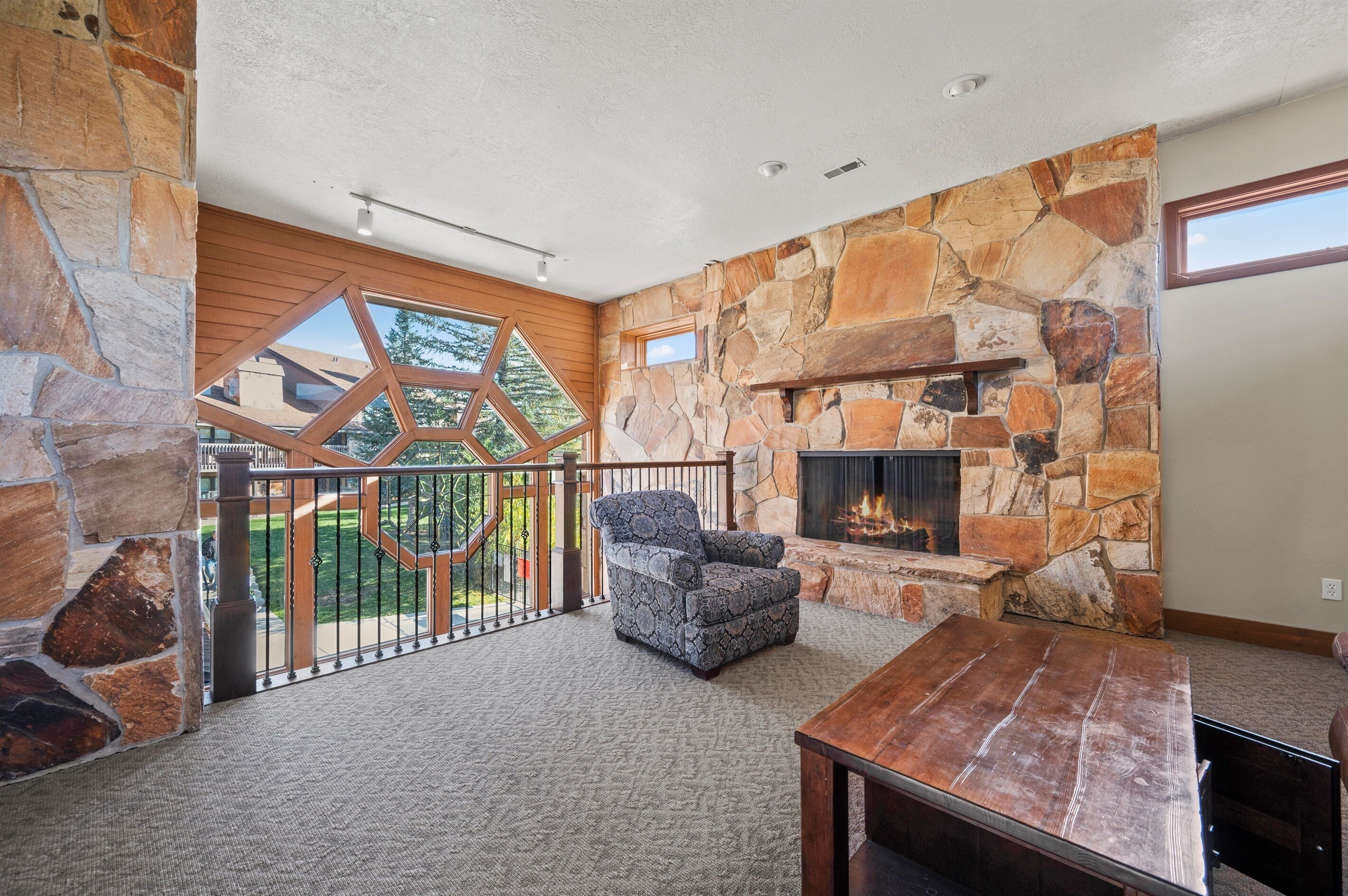 A spacious living area with a stone fireplace, a wooden bench, and a view of a landscaped deck.