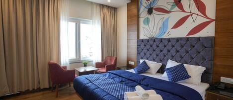 A hotel room with a large bed, a headboard, a desk, a chair, and a window with curtains.