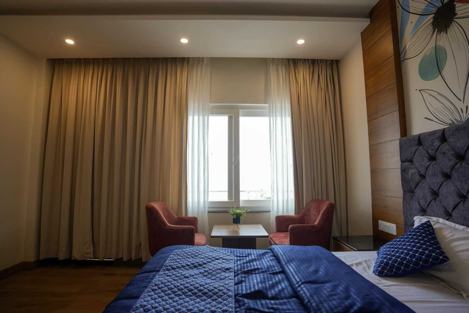 A hotel room with a bed, two red armchairs, a small table, and a window with curtains.